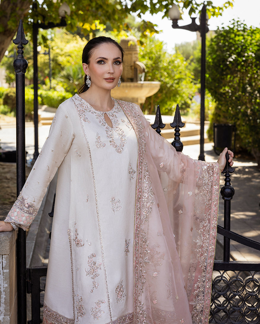 Woman wearing Astrid Bloom off white embroidered raw silk suit with pink organza dupatta at garden gate
