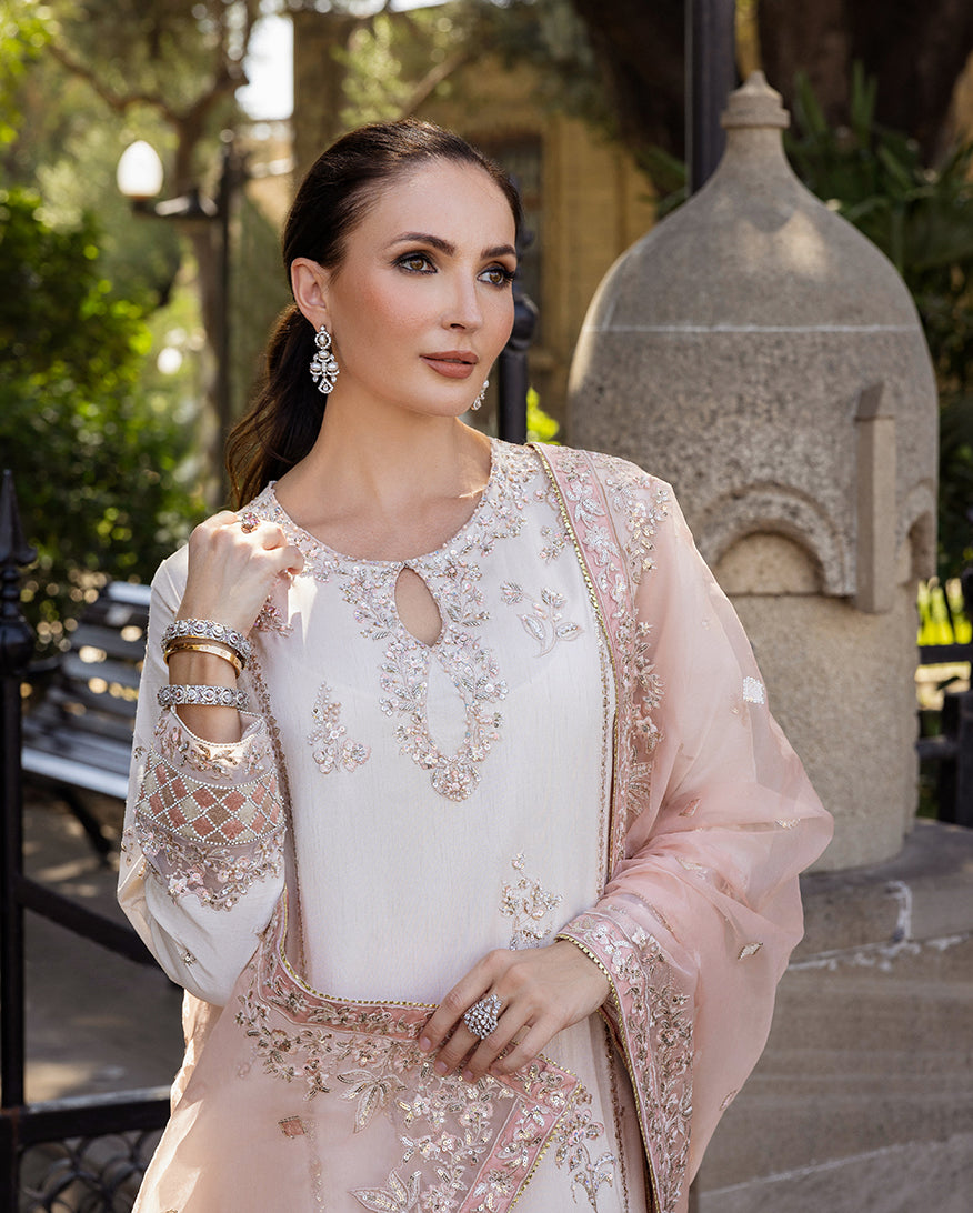 Woman wearing Astrid Bloom off white embroidered raw silk suit with pink organza dupatta at garden gate