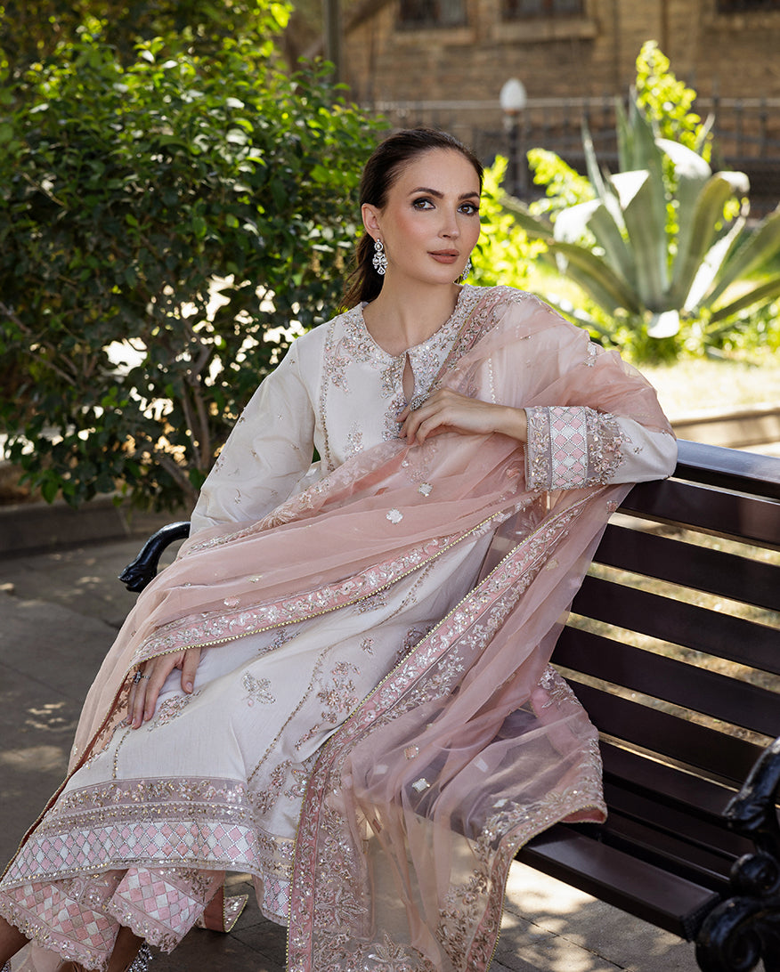 Woman wearing Astrid Bloom off white embroidered raw silk suit with pink organza dupatta at garden gate