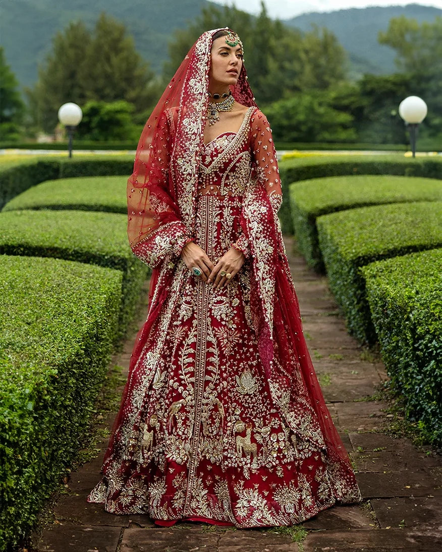Woman wearing blood red embellished Net bridal lehenga with sheer veil, standing in garden