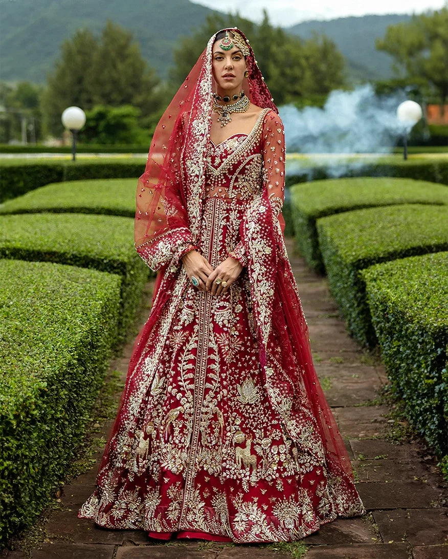 Woman wearing blood red embellished Net bridal lehenga with sheer veil, standing outdoors