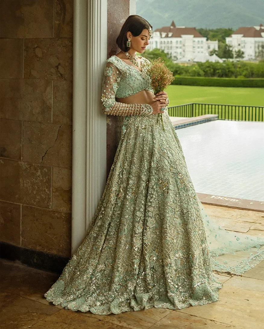 Woman wearing aqua green embellished Net bridal lehenga, leaning against pillar with bouquet