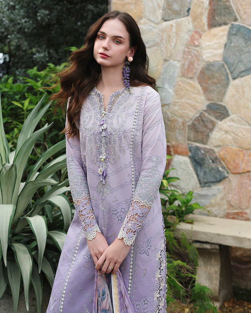 Woman wearing lavender lawn unstitched collection suit with lace sleeve trim and tassel necklace, standing near stone wall, Secret Garden collection