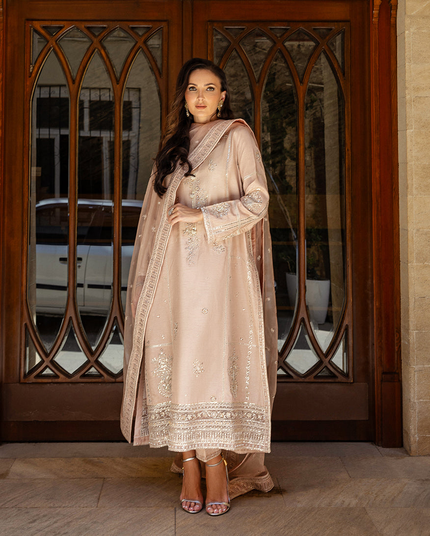 Woman wearing rose beige embroidered Korean raw silk suit with sheer organza dupatta at arched wooden doorway
