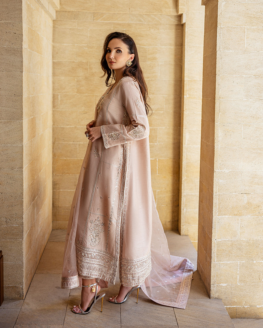 Side view of rose beige raw silk suit with organza dupatta draped over arm in stone corridor